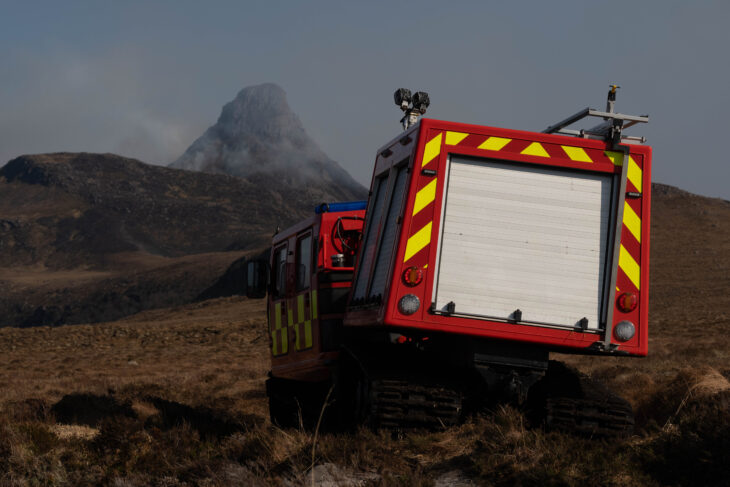 A red all terrain vehicle driving over rough ground towards a smoking mountain.